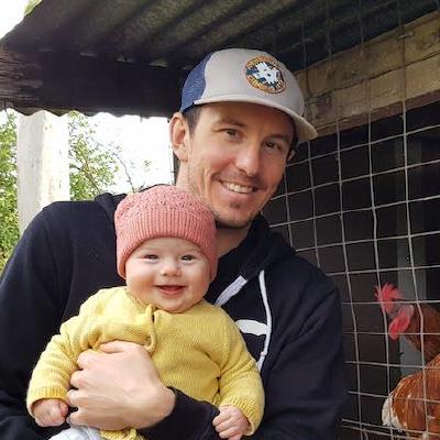 Jonathan smiling with a baby in a pink beanie, in front of a chicken coop